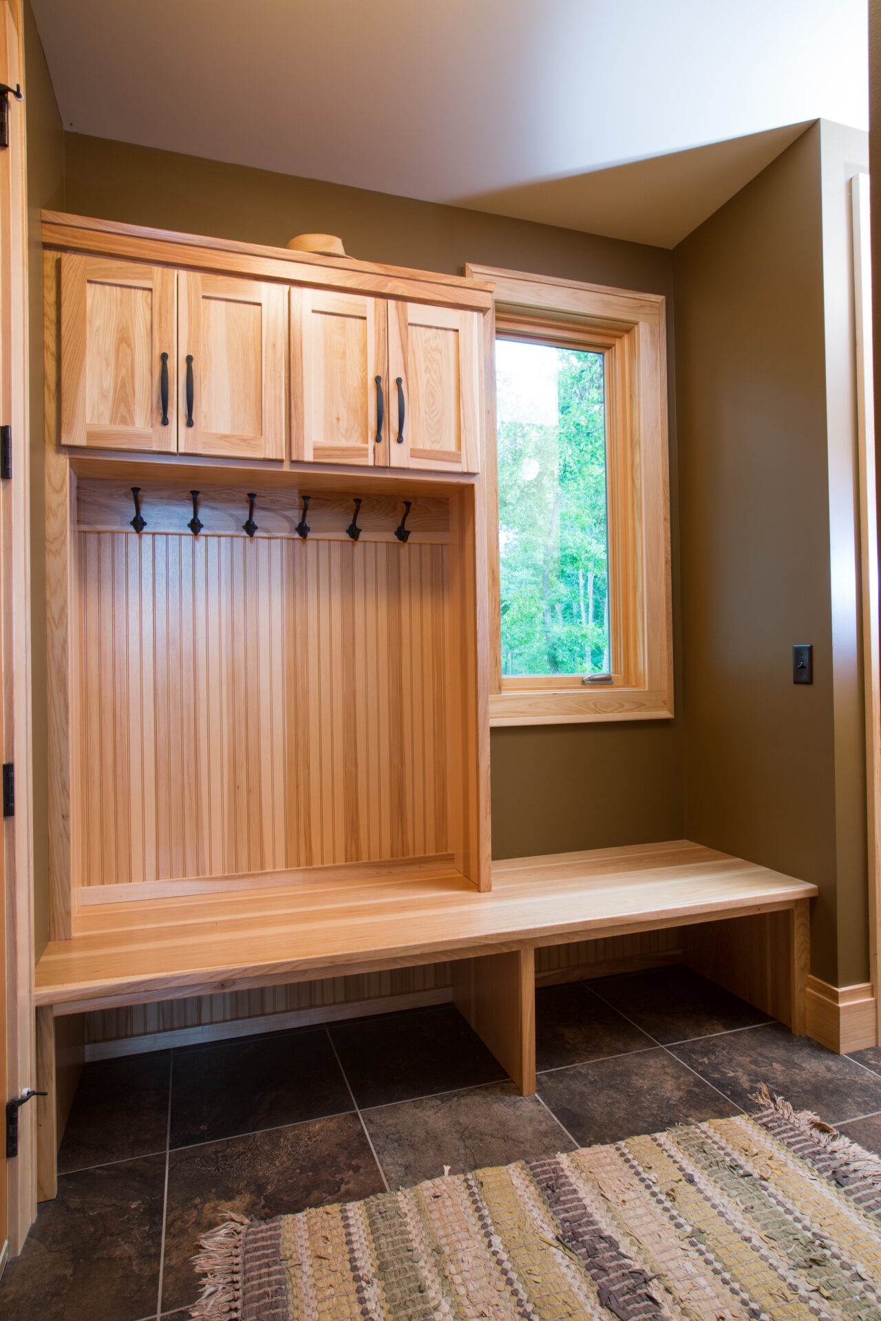 A wooden bench with hooks and cabinets in front of a window.